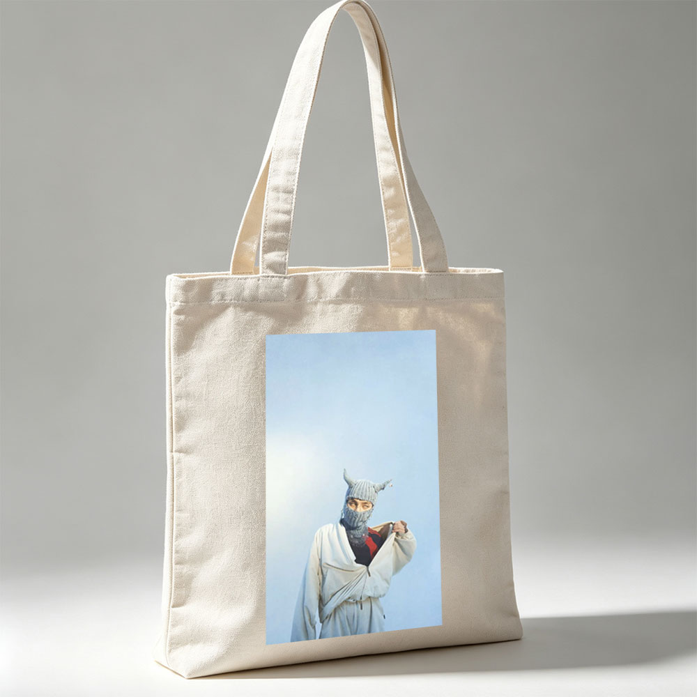 Billie Eilish in a whimsical winter scene wearing a horned knit mask and flowing white outfit against a clear blue sky - Canvas tote bags