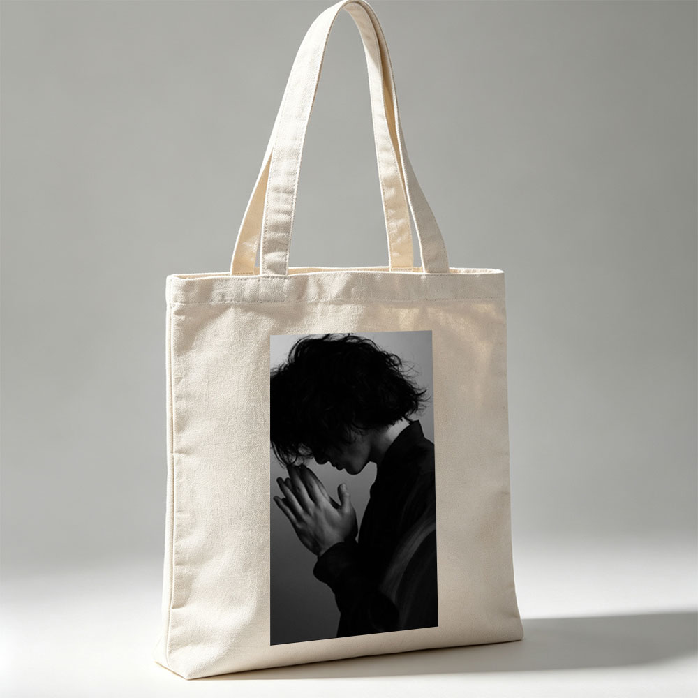 Yojiro Noda in a contemplative pose with hands clasped, captured in dramatic black and white lighting. - Canvas tote bags