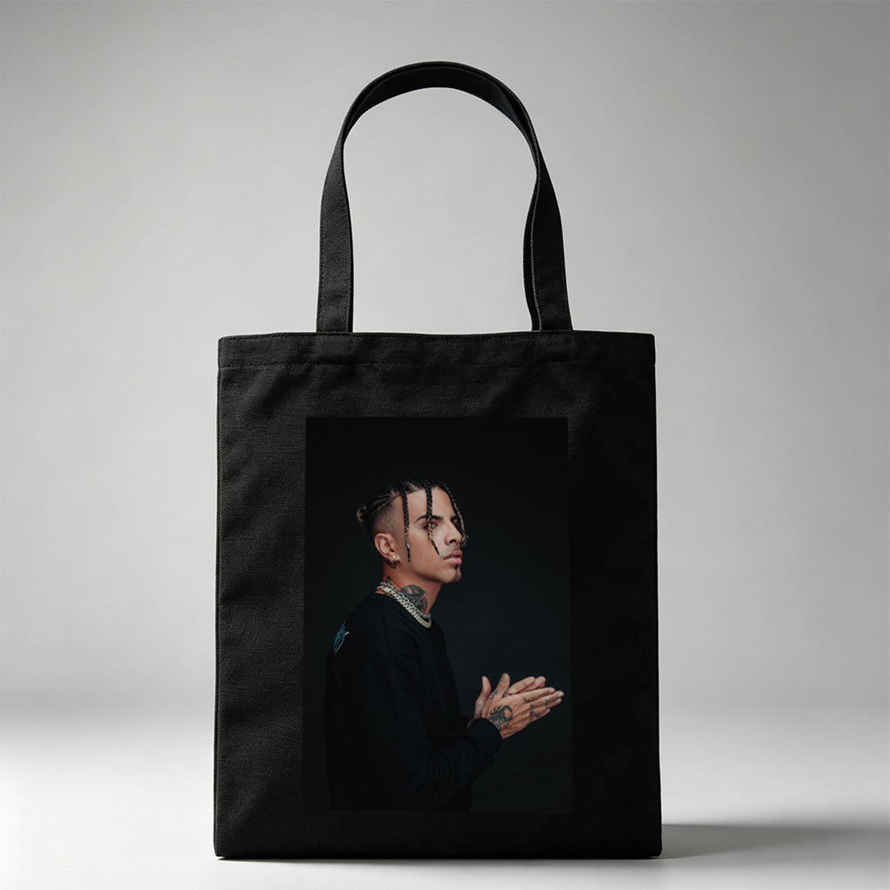 Daddy Yankee in a striking pose with braided hair and tattoos, wearing a black outfit and gold chain against a dark background. - Canvas tote bags