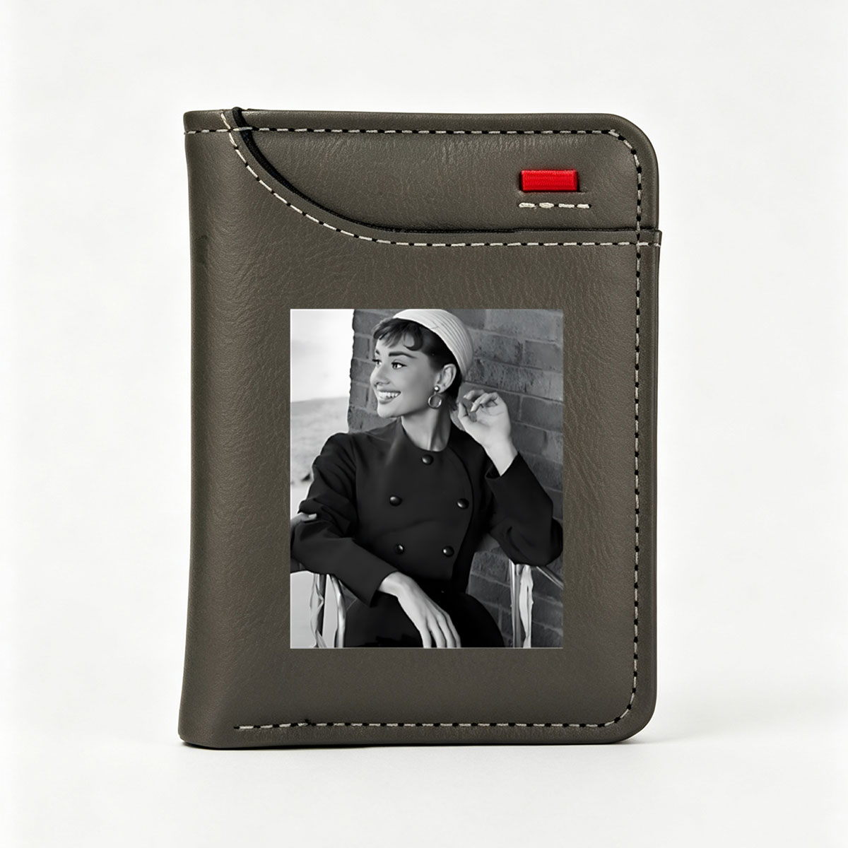 Audrey Hepburn in a classic black coat and white hat, smiling elegantly against a brick wall. - Vertical Style Wallet