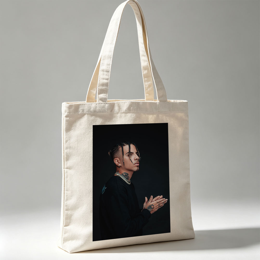Daddy Yankee in a striking pose with braided hair and tattoos, wearing a black outfit and gold chain against a dark background. - Canvas tote bags