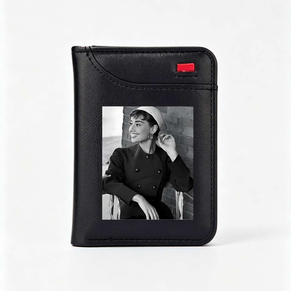 Audrey Hepburn in a classic black coat and white hat, smiling elegantly against a brick wall. - Vertical Style Wallet