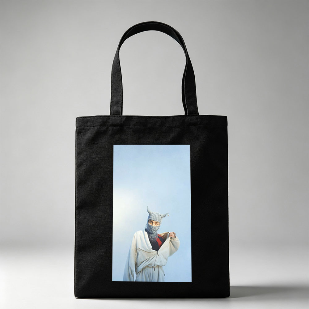 Billie Eilish in a whimsical winter scene wearing a horned knit mask and flowing white outfit against a clear blue sky - Canvas tote bags